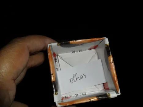 Fotografia em fundo preto de mão segurando caixa de papel, dentro dela um papel com a palavra olhos